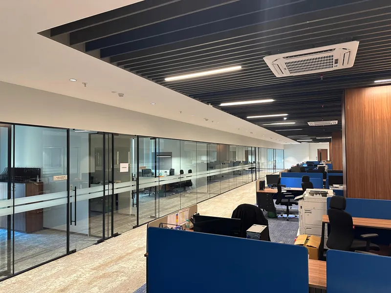 Open-plan office with glass-partitioned cabins and dark baffle ceiling with linear lighting.