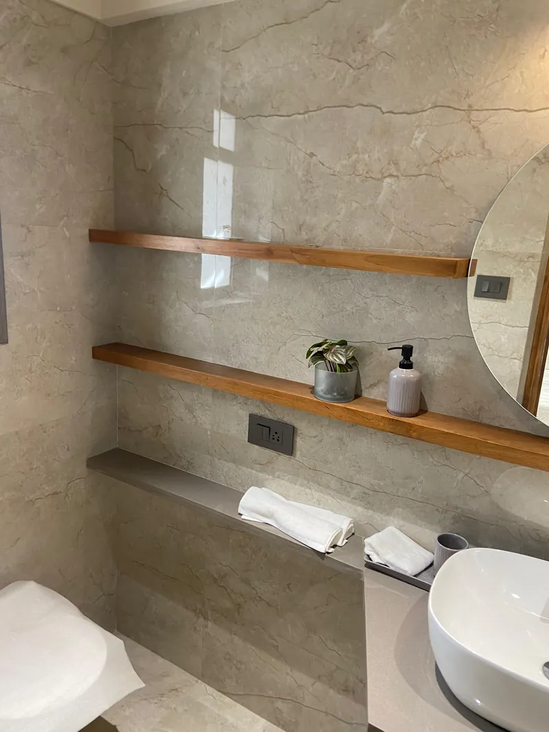 Marble bathroom with wooden floating shelves, round mirror and vessel sink.