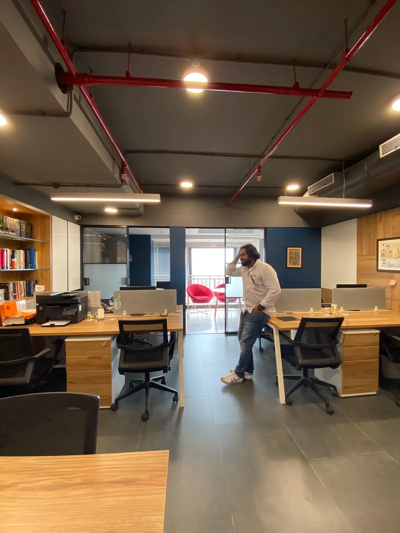 Office with clustered wood desks, mesh chairs and exposed red ceiling piping.